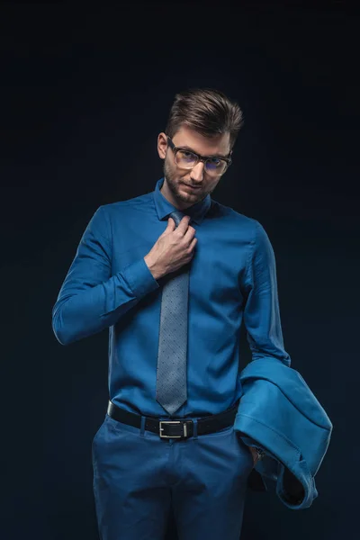 Joven hombre de negocios en gafas fijando su corbata aislada en azul - foto de stock