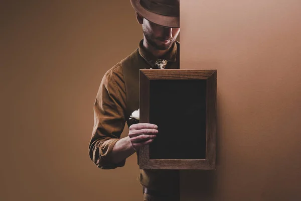 Stylish man in hat holding empty frame isolated on brown — Stock Photo