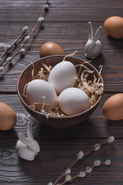 Uova di pollo in ciotola con paglia e conigli pasquali sul tavolo di legno — Foto stock