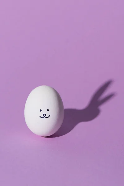 Un œuf de poulet avec un sourire et une ombre de lapin sur la surface violette — Photo de stock