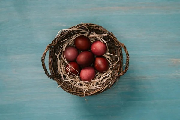 Верхний вид пасхальной корзины с покрашенными яйцами и соломой на столе — стоковое фото