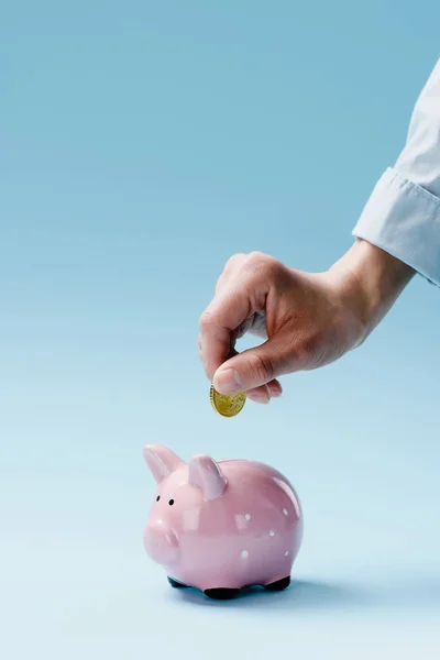 Vista parcial del hombre poniendo moneda en la alcancía rosa aislado en azul - foto de stock