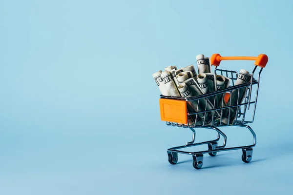 Close up view of little shopping trolley with rolled dollar banknotes isolated on blue — Stock Photo