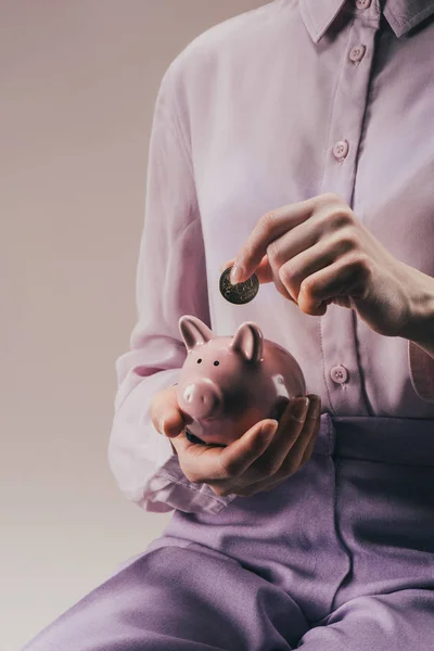 Cropped shot of woman putting 50 euro cent into pink piggy bank in hand isolated on lilac — Stock Photo