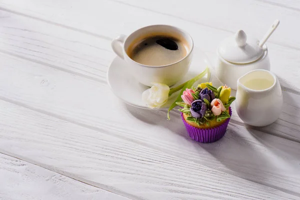 Vista de perto da xícara de café, flor de tulipa, muffin doce e jag de creme na mesa de madeira branca — Fotografia de Stock