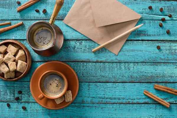 Flat lay com xícara de café, tigela com açúcar mascavo, envelope, grãos de café torrados e paus de canela em torno de mesa de madeira azul — Fotografia de Stock