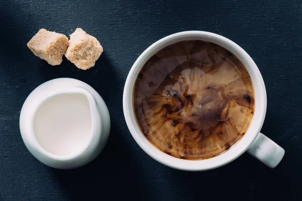 Flat lay com xícara de café, jag de creme e pedaços de açúcar de cana na superfície escura — Fotografia de Stock