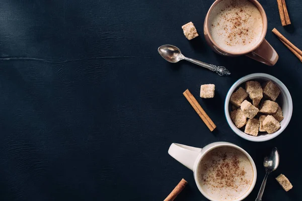 Top view of cups of coffee, spoons, bowl of cane sugar and cinnamon stick on dark tabletop — Stock Photo