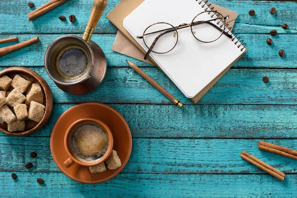 Flat lay with cup of coffee, eyeglasses, bowl with brown sugar, empty notebook, roasted coffee beans and cinnamon sticks around on blue wooden tabletop — Stock Photo