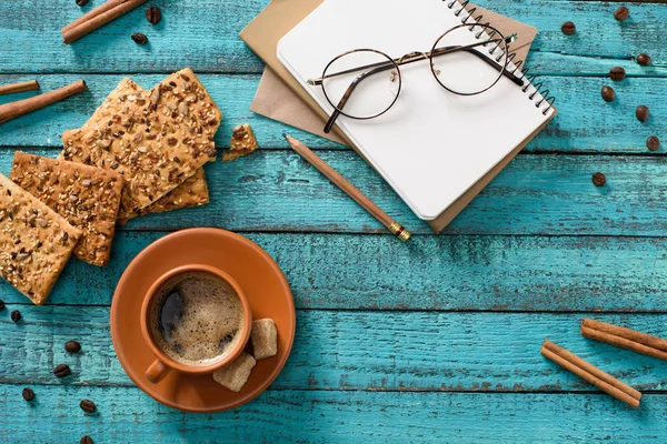 Flat lay com xícara de café, biscoitos, óculos, caderno vazio, grãos de café torrados e paus de canela em torno de mesa de madeira azul — Fotografia de Stock