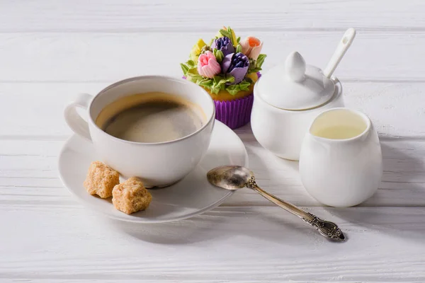 Close up view of cup of coffee, sweet muffin, spoon and jag of cream on white wooden tabletop — Stock Photo