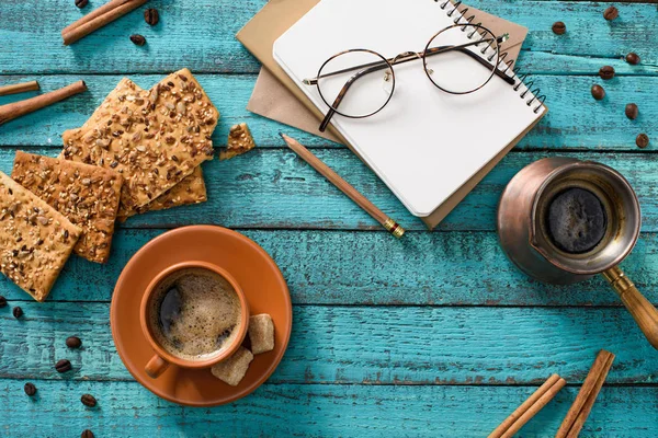 Posa piatta con tazza di caffè, biscotti, occhiali, quaderno vuoto, chicchi di caffè torrefatto e bastoncini di cannella intorno su un tavolo di legno blu — Foto stock