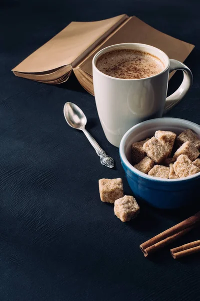 Cerrar vista del libro, taza de café, tazón con azúcar morena y palitos de canela en la superficie oscura - foto de stock