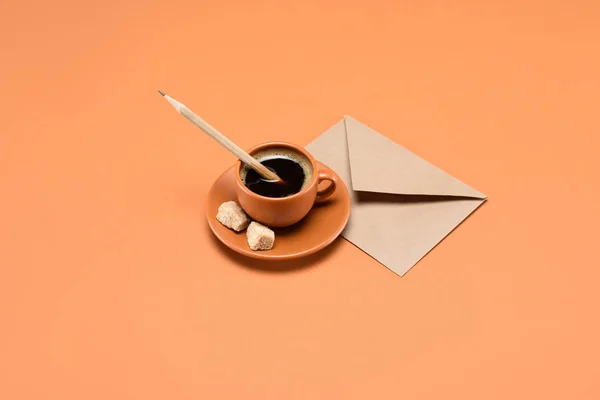 Close up view of cup of coffee with pencil, cane sugar on saucer and envelope near by isolated on peach — Stock Photo