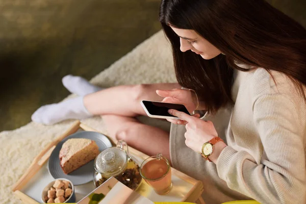 Alto angolo di vista della donna con smartphone e tazza di tè su vassoio di legno a casa — Foto stock