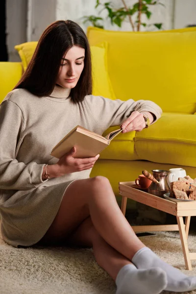 Livre de lecture femme assis sur le sol près d'un plateau en bois avec tasse de café et de pâtisserie à la maison — Photo de stock
