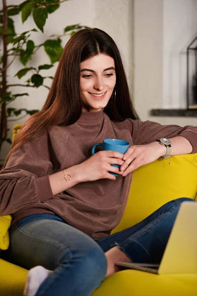 Женщина с чашкой кофе с помощью ноутбука на диване дома — стоковое фото