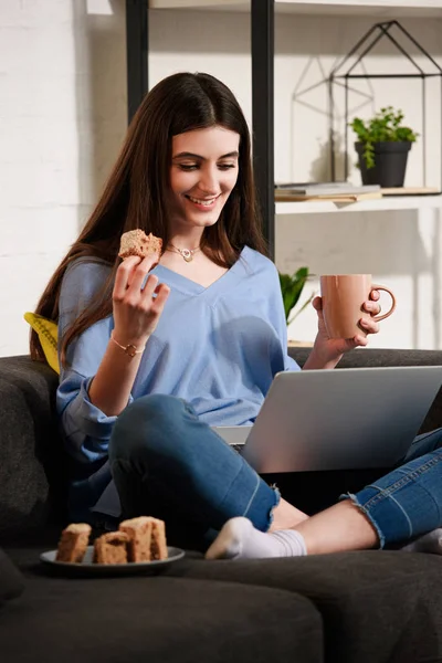 Улыбающаяся женщина с чашкой кофе и кусок торта с помощью ноутбука на диване дома — стоковое фото