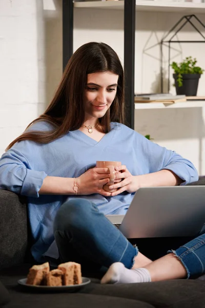Donna con tazza di caffè utilizzando il computer portatile sul divano di casa — Foto stock