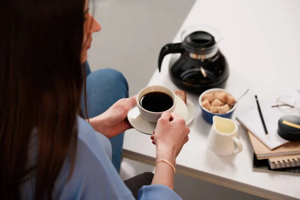 Partial view of woman with cup of coffee and coffee table with coffee maker, jag of cream and brown sugar — Stock Photo