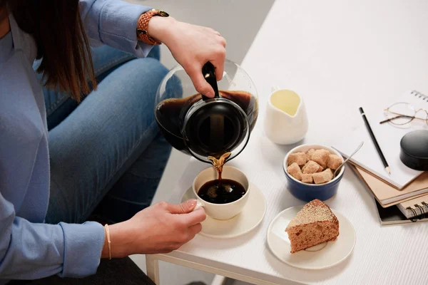 Tiro recortado de la mujer vertiendo café en la taza con azúcar de caña y pedazo de pastel en la mesa de café - foto de stock