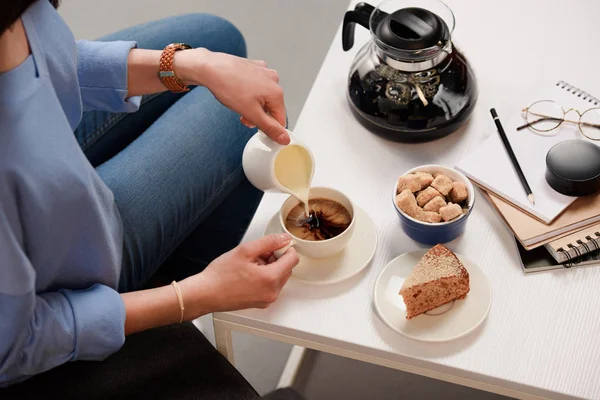 Tiro recortado de la mujer verter crema en la taza de café con azúcar de caña y pedazo de pastel en la mesa de café - foto de stock