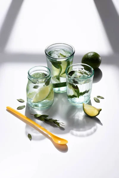 Nahaufnahme von Plastiklöffel, Gläsern mit Wasser, Limettenstücken und Eiswürfeln auf weißer Tischplatte — Stockfoto