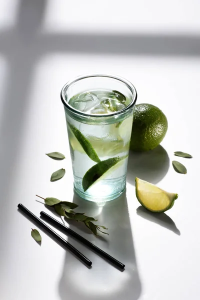 Nahaufnahme von Strohhalmen, einem Glas Wasser mit Limettenstücken und Eis auf einer weißen Tischplatte — Stockfoto