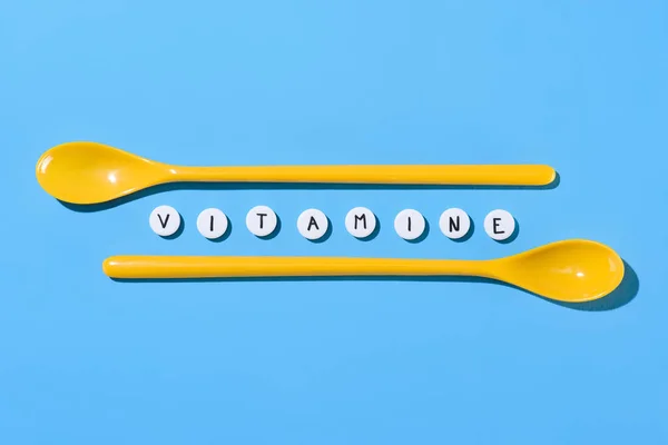 Top view of yellow plastic spoons and pills with vitamine lettering on blue — Stock Photo