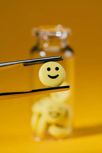 Close-up shot of pill with smiley face in tweezers and glass bottle on yellow — Stock Photo