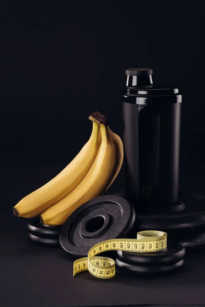 Black protein shaker on pile of weight plates with bananas and measuring tape isolated on black — Stock Photo