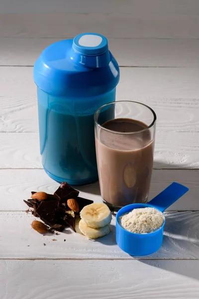 Agitador e copo de batido de proteína com amêndoa, banana e chocolate na mesa de madeira branca — Fotografia de Stock