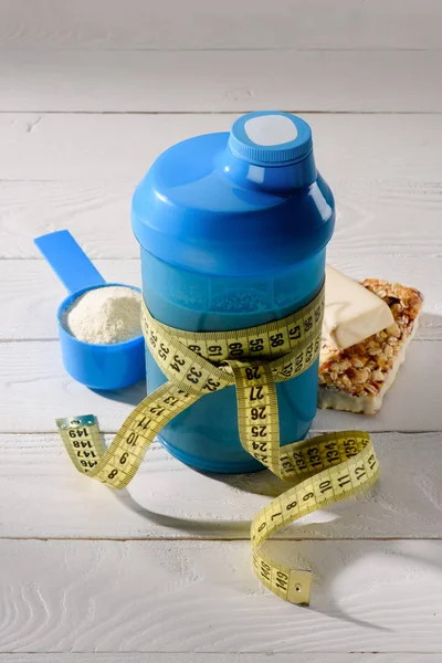 Close-up shot of protein shaker tied with measuring tape and energy bars on white wooden table — Stock Photo