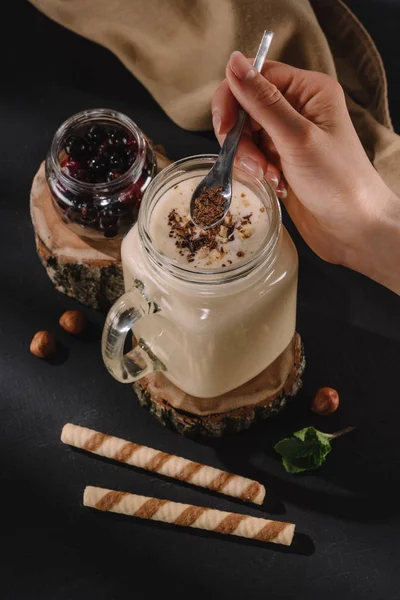 Tiro cortado da mão feminina colocando raspas de chocolate em milkshake, hortelã, palhas doces e groselhas na mesa — Fotografia de Stock
