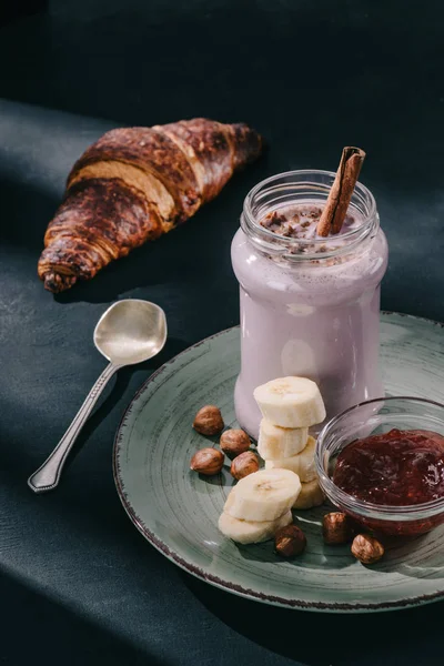 Primer plano de batido de bayas con paja de canela y virutas de chocolate, nueces, mermelada y rodajas de plátano en el plato, cuchara y croissant - foto de stock