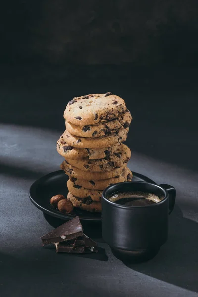 Pila de galletas de chispas de chocolate en el plato con taza de café en la superficie negra - foto de stock