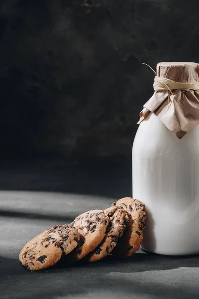 Delicious chocolate-chip cookies with glass bottle of milk on black surface — Stock Photo