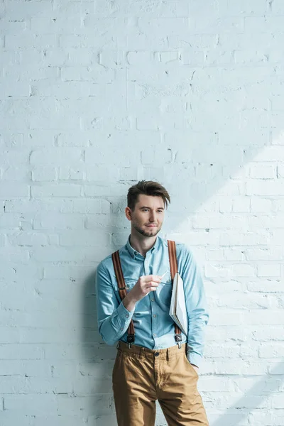 Retrato de arquiteto pensativo com lápis e notebook olhando para longe enquanto de pé contra a parede de tijolo branco — Fotografia de Stock