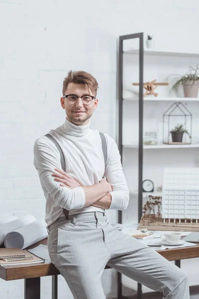 Portrait of smiling architect in eyeglasses with arms crossed leaning on workplace in office — Stock Photo