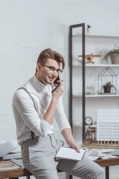 Portrait of architect in eyeglasses with notebook talking on smartphone at workplace in office — Stock Photo