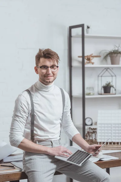 Portrait of smiling architect in eyeglasses with laptop leaning on workplace in office — Stock Photo