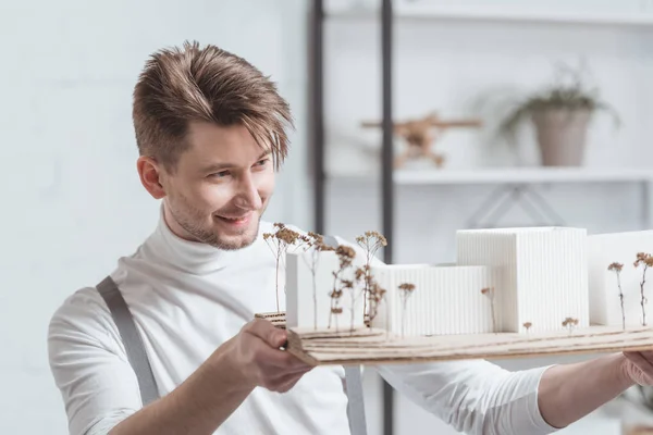 Портрет улыбающегося архитектора, смотрящего на самодельную модель здания в руках в офисе — стоковое фото