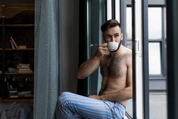 Мужчина без рубашки пьет кофе на подоконнике — Бесплатное стоковое фото
