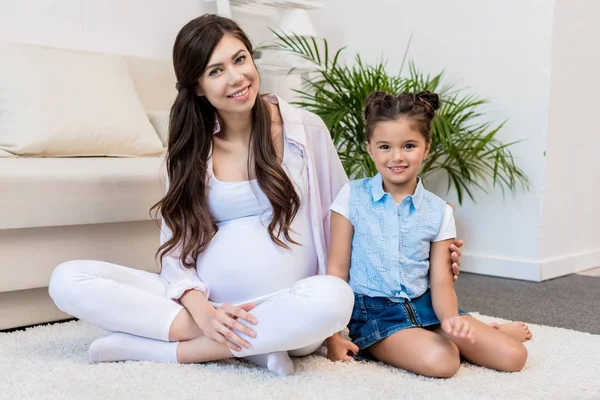 Zwangere vrouw met dochter zit op tapijt — Stockfoto