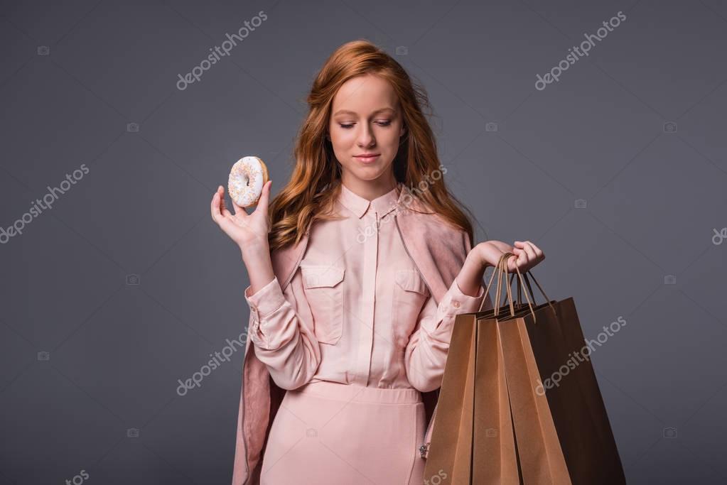 Attractive elegant lady in pink clothes with donut and shopping bags, isolated on grey