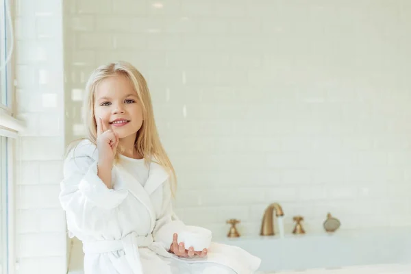 Niño en albornoz aplicando crema facial — Foto de Stock