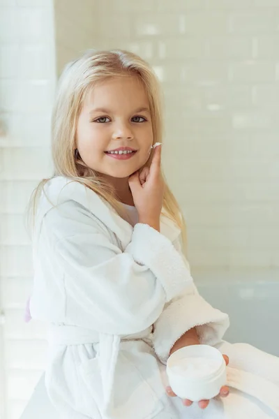 Child in bathrobe applying face cream — Stock Photo, Image