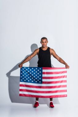 athletic african american sportsman holding american flag and looking at camera on grey clipart