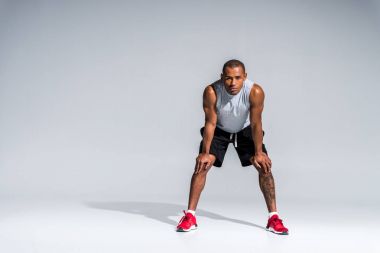 elleriyle dizlerinin üzerinde duran ve gri kameraya bakarak atletik genç Afro-Amerikan sporcu görünümünü tam uzunlukta