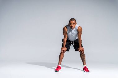 full length view of athletic young african american sportsman standing with hands on knees and looking at camera on grey clipart